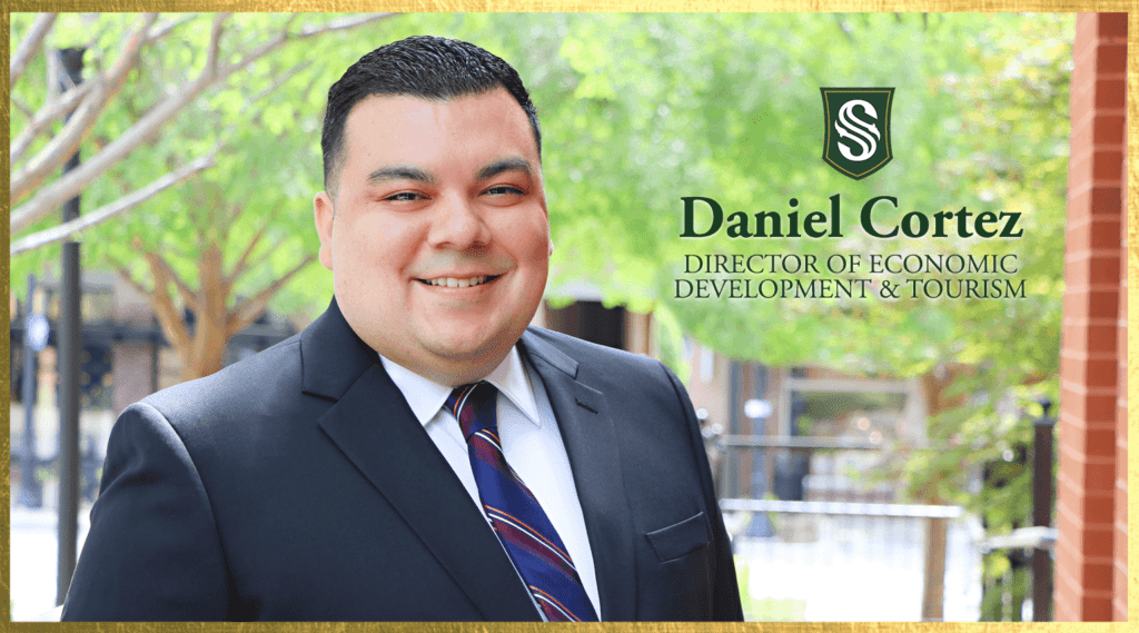 A man in a suit and tie smiles outdoors. Text next to him reads "Daniel Cortez, Director of Economic Development & Tourism." Green trees and a blurred background add a natural setting.